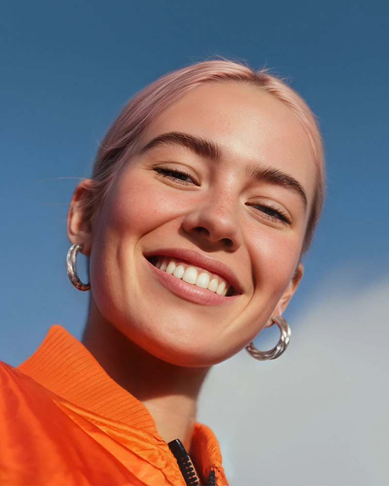 Junge Frau mit blonden Haaren und Ohrringen, lachend vor blauem Himmel.