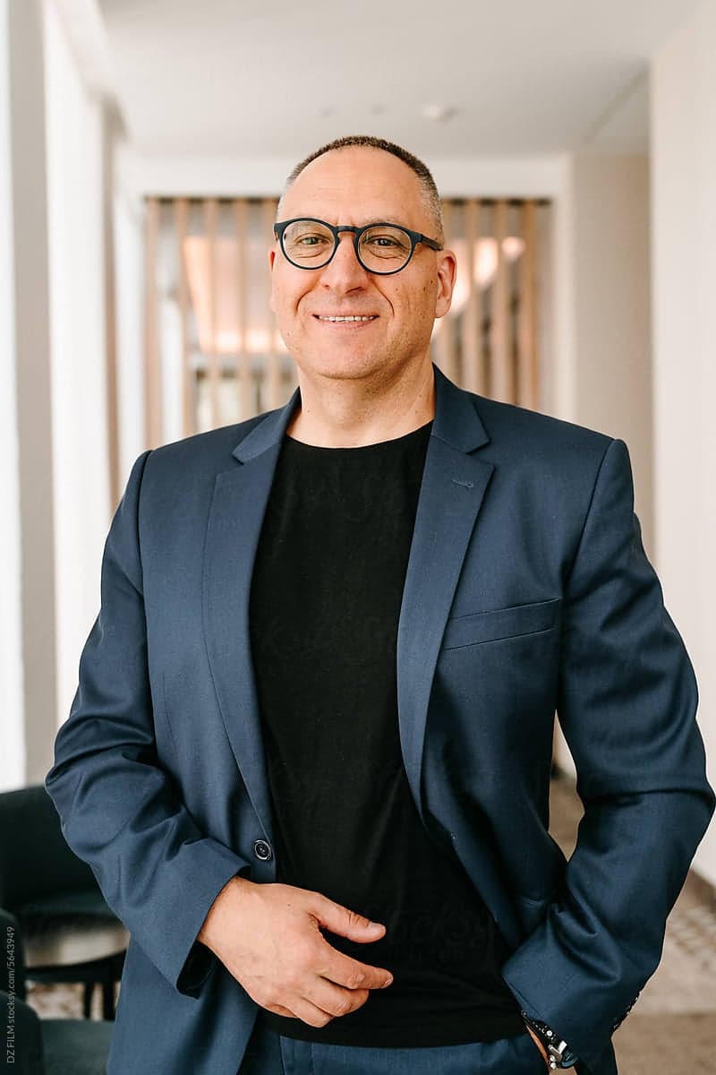 A respectable man with glasses, dressed in a business suit, smiles looking at the camera standing in a stylish office