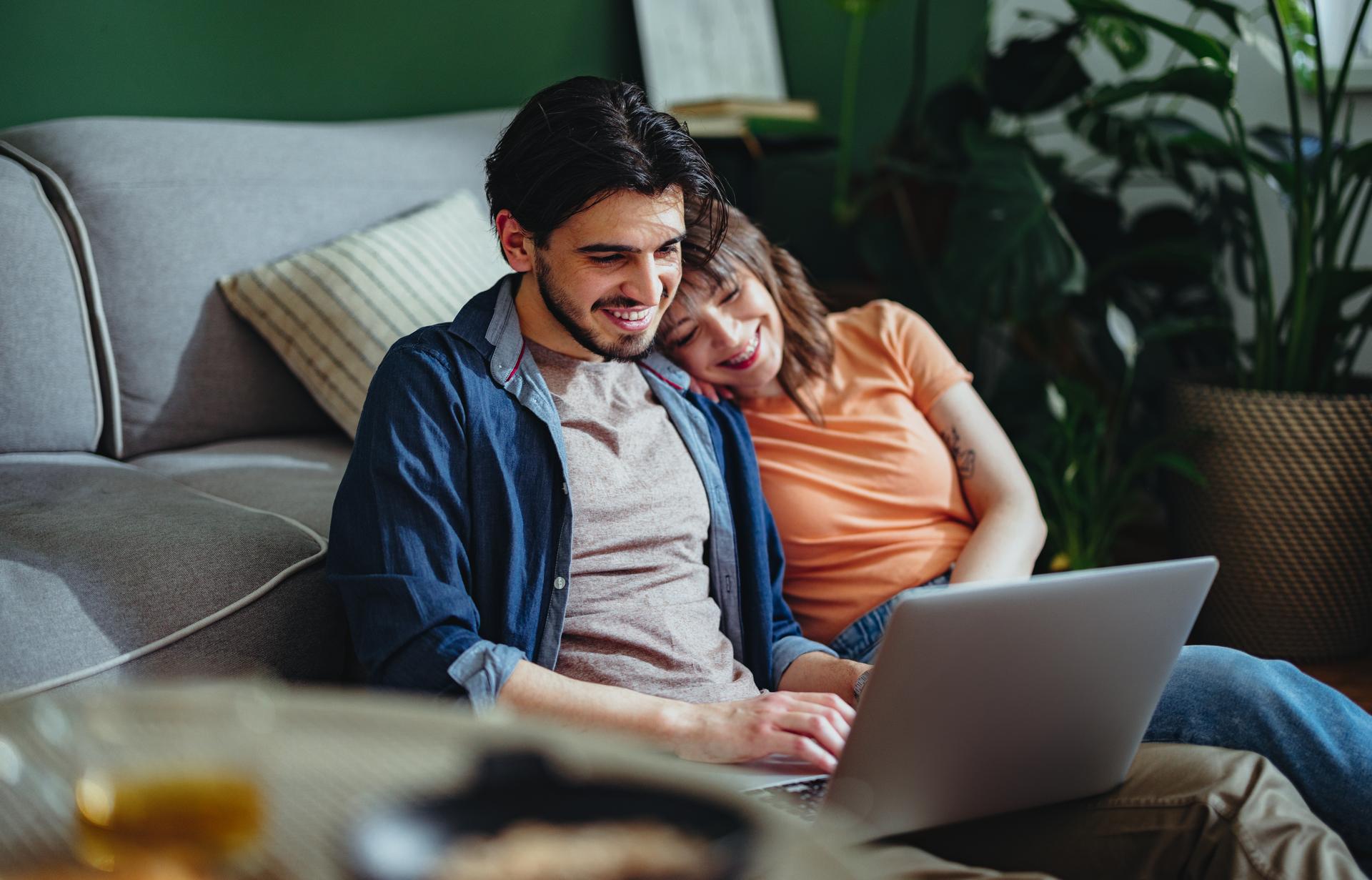 Cheerful boyfriend typing something on a laptop keyboard while sitting on the floor in the living room with his girlfriend.
