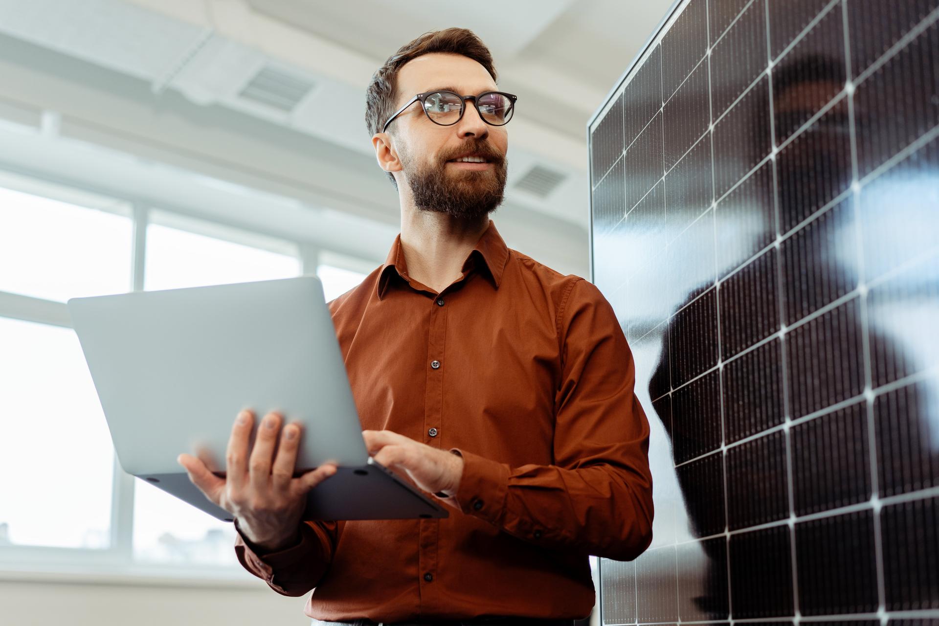 Handsome smiling man, engineer, developer holding laptop computer planning startup working with solar panel in modern office. Innovation, renewable energy concept