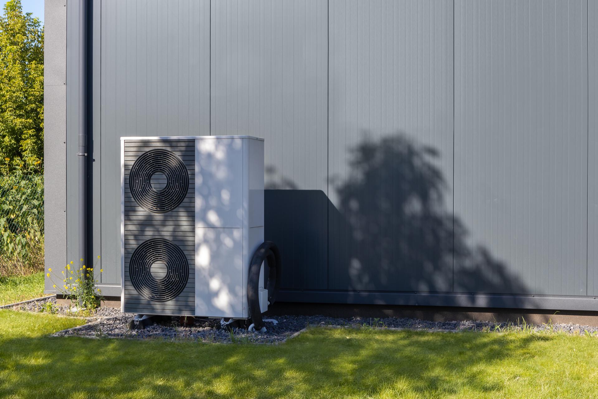 Sleek white heat pump with dual fans installed beside a gray industrial building, surrounded by grass and tree shadows on a sunny day.