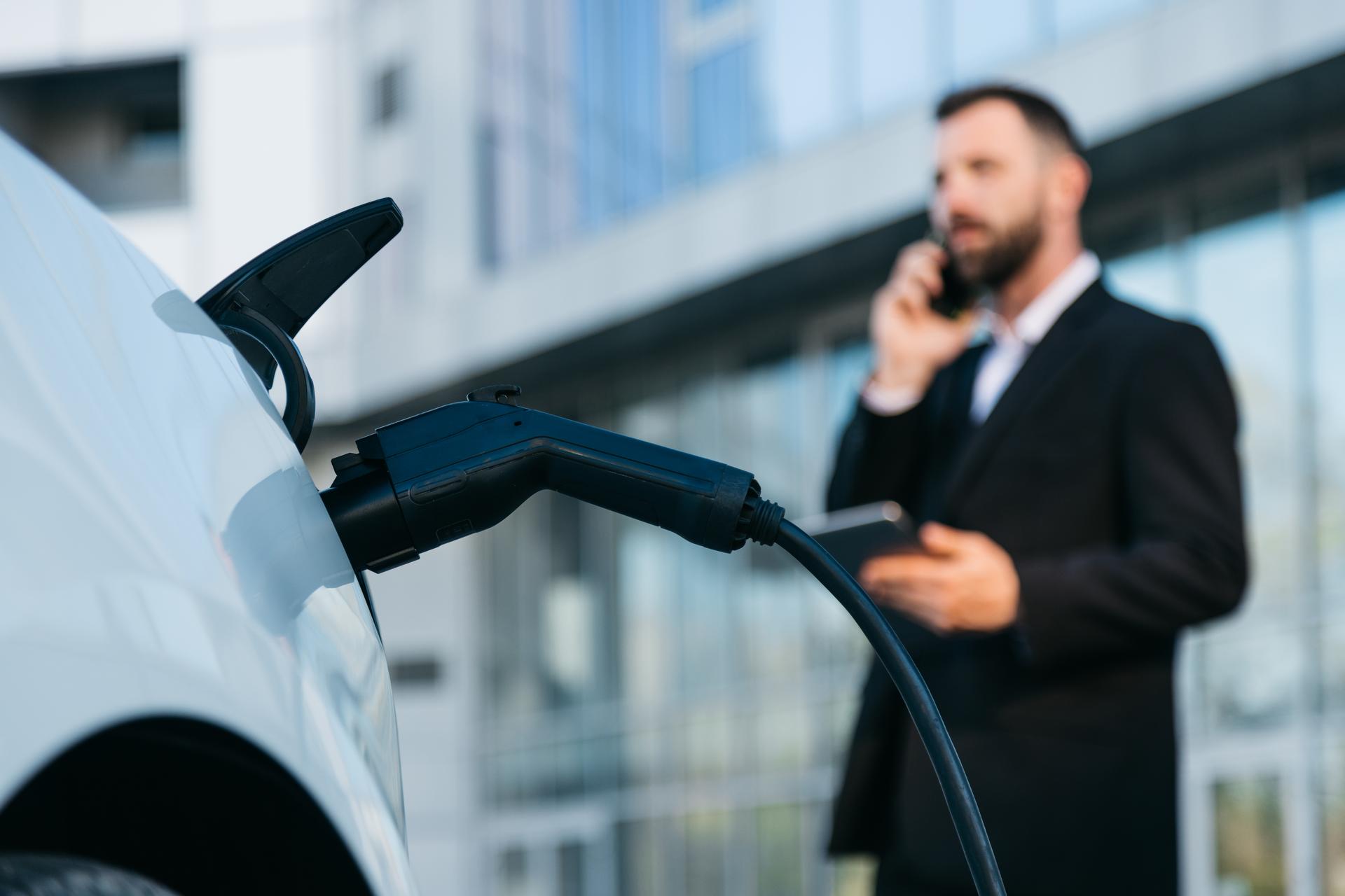 A business man talking on his cell phone while charging the battery of his electric car. A confident man in a business suit near a luxury car. Eco-friendly transportation in an urban environment.