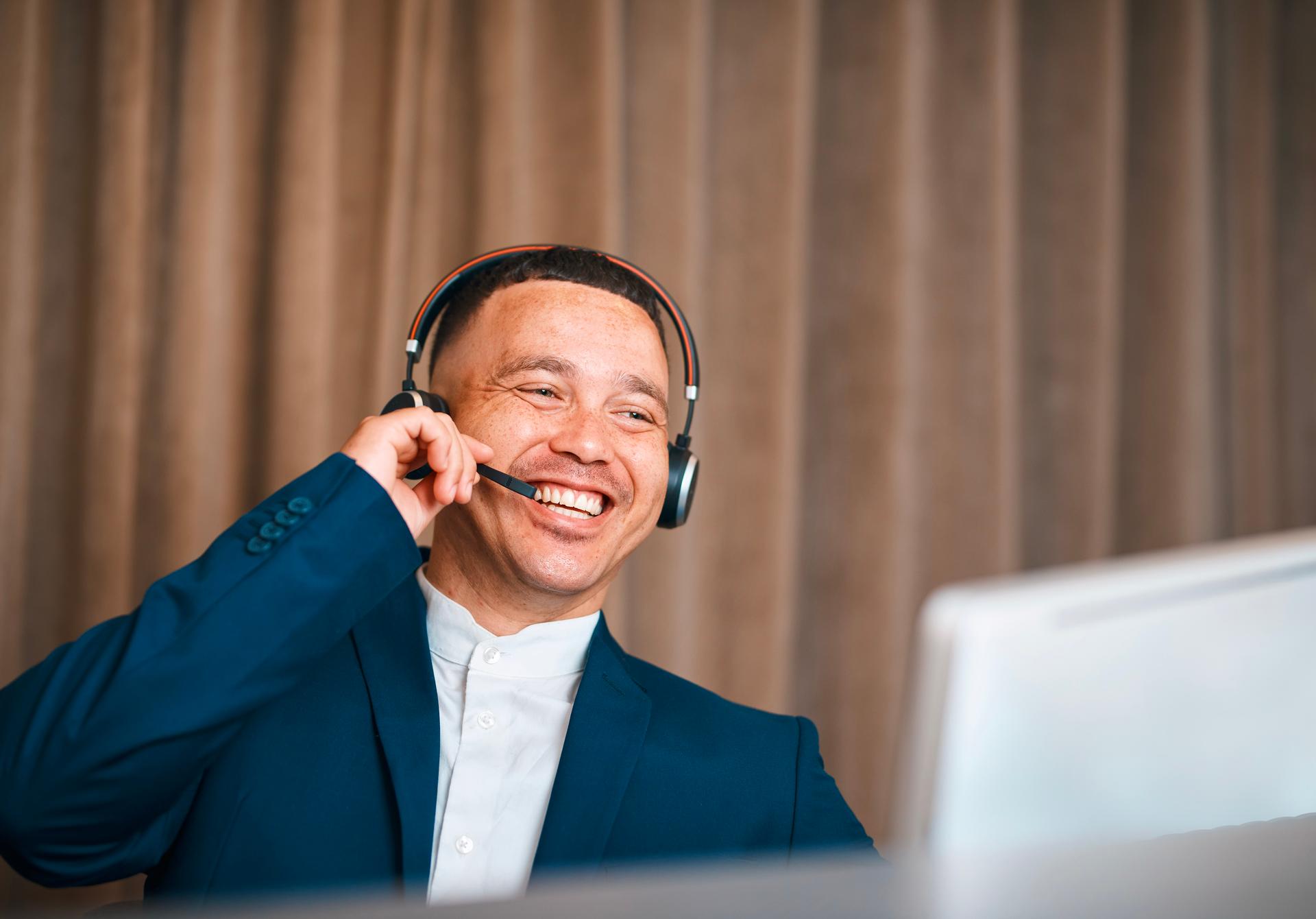 Friendly professional in a blue suit with a headset, engaging with a computer at a reception or desk, conveying customer service, support, and efficient hospitality in a corporate setting.