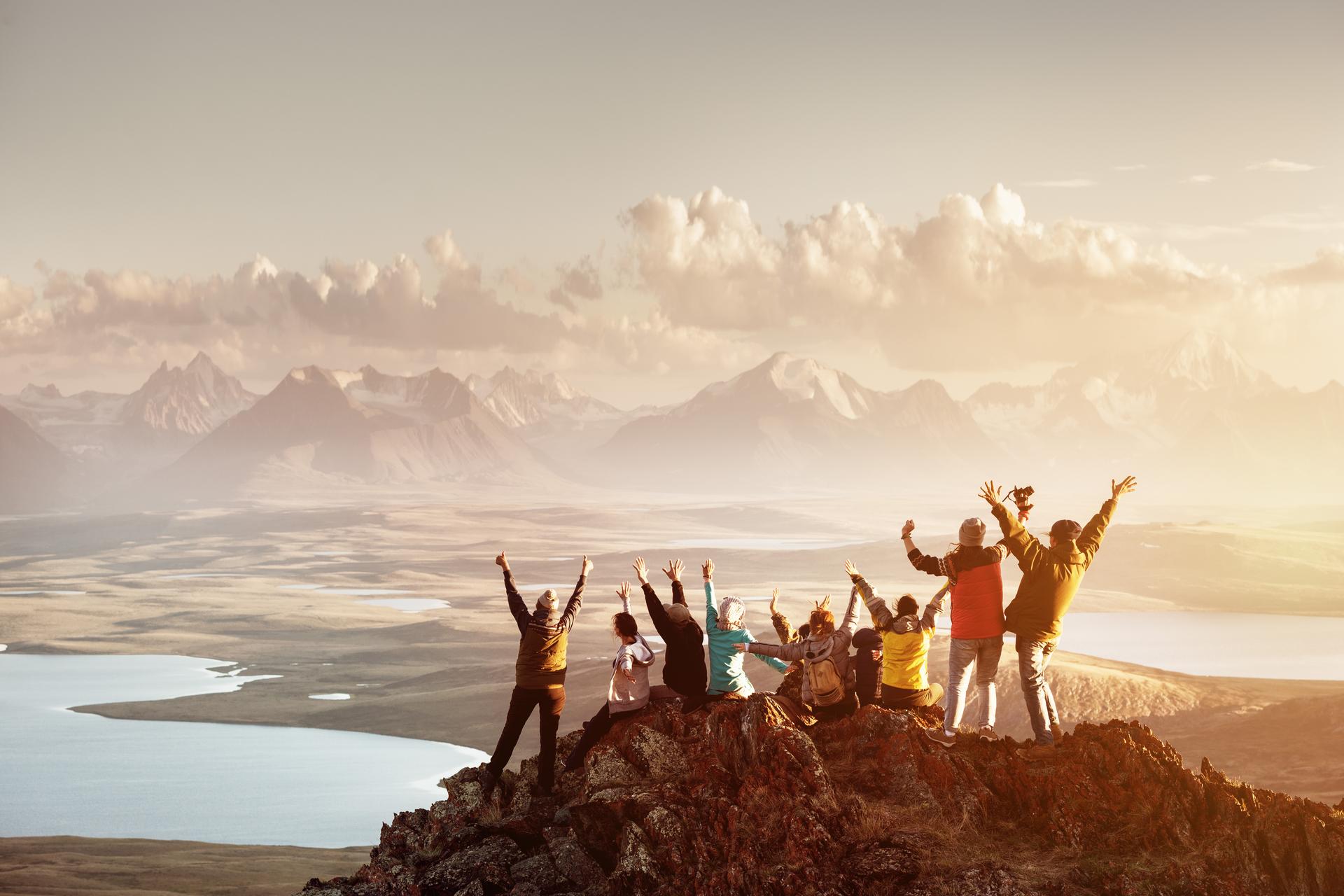 Big group of people having fun in success pose with raised arms on mountain top against sunset lakes and mountains. Travel, adventure or expedition concept