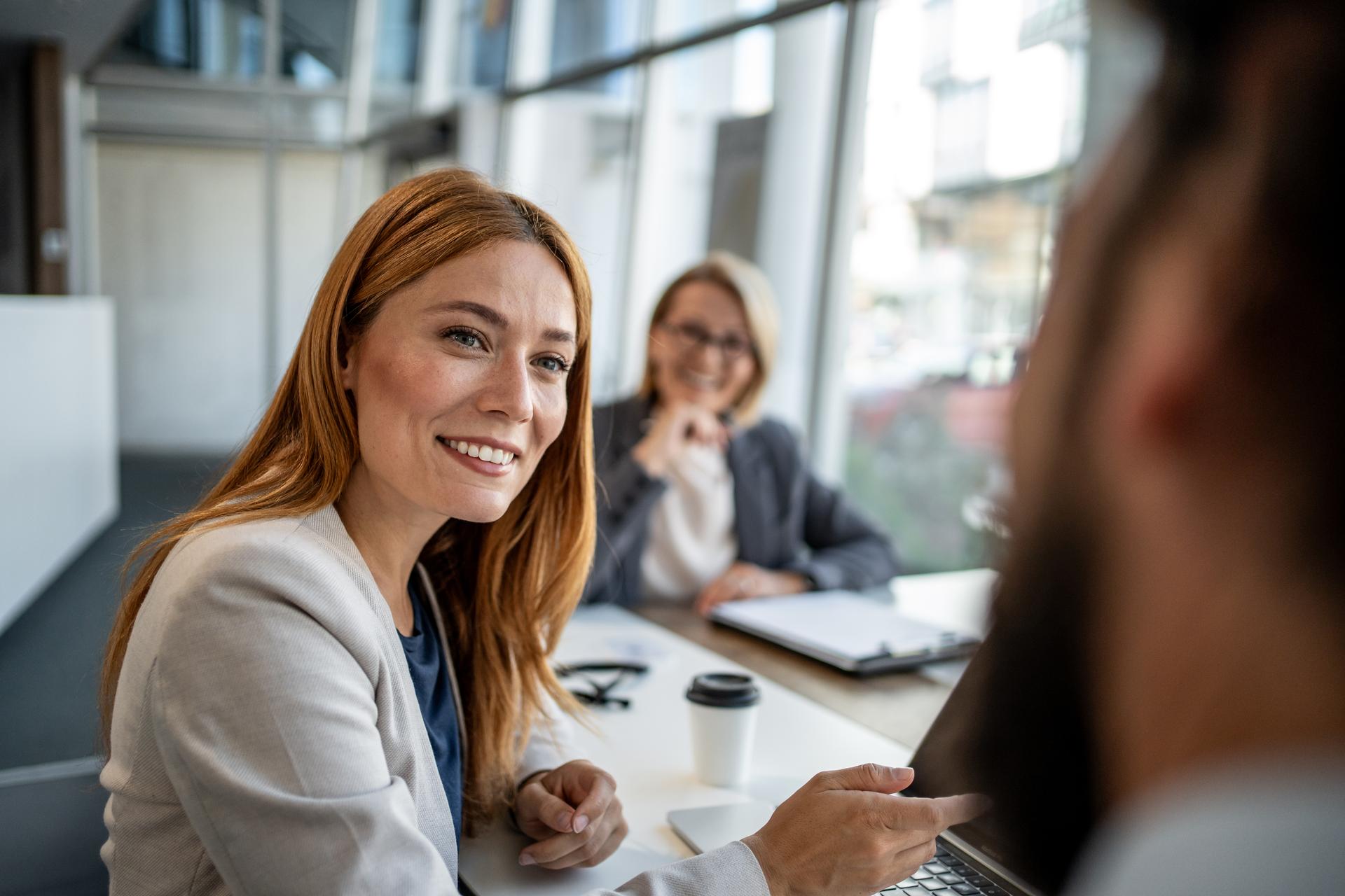Businesswomen smiling and engaging in a lively discussion about a project during a collaborative meeting with colleagues in a modern office setting, showcasing teamwork and professionalism